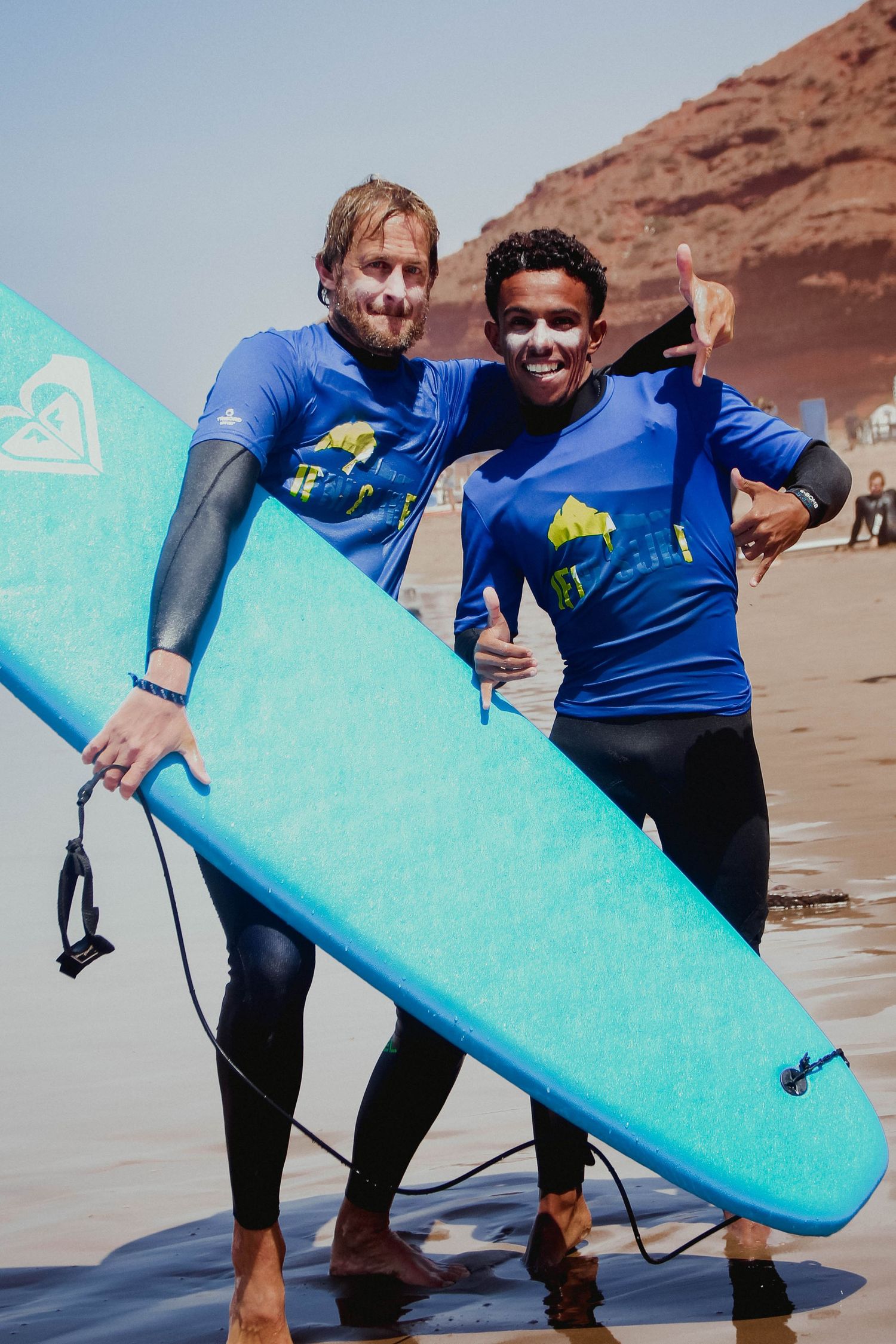 Two smiling surfers in blue rash guards holding a turquoise surfboard on a sandy beach with red cliffs.