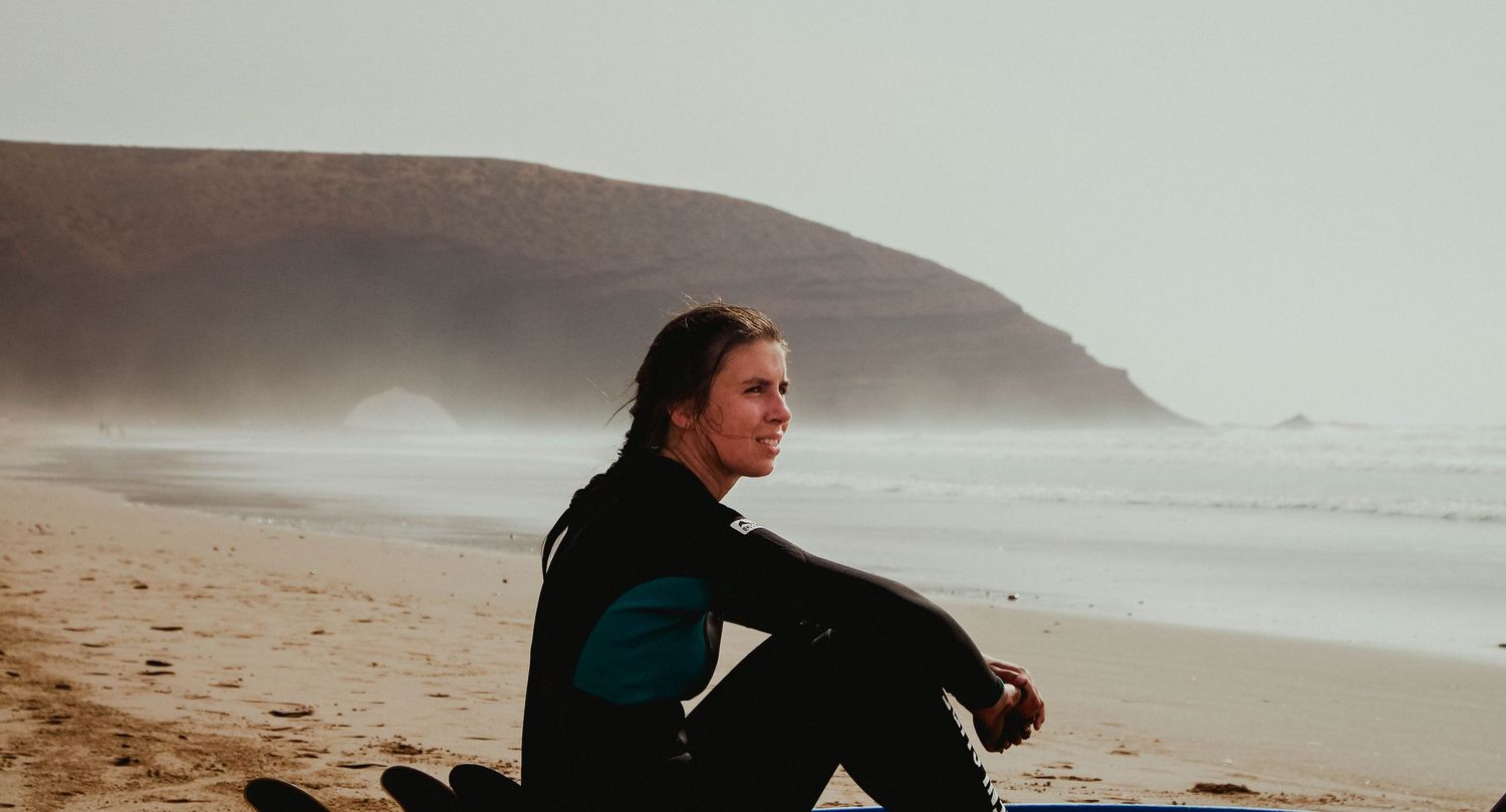 A woman in a wetsuit sits on a sandy beach, gazing thoughtfully at misty coastal cliffs in the background.