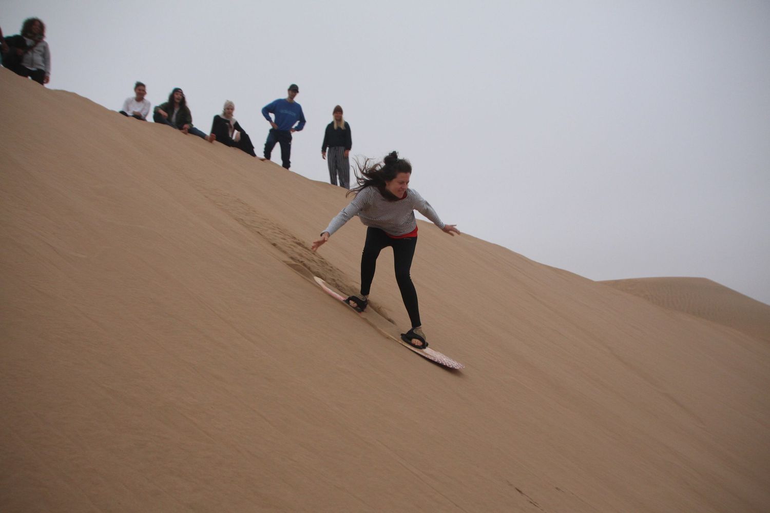Person sandboarding down a steep sand dune in a desert landscape with spectators watching from above.