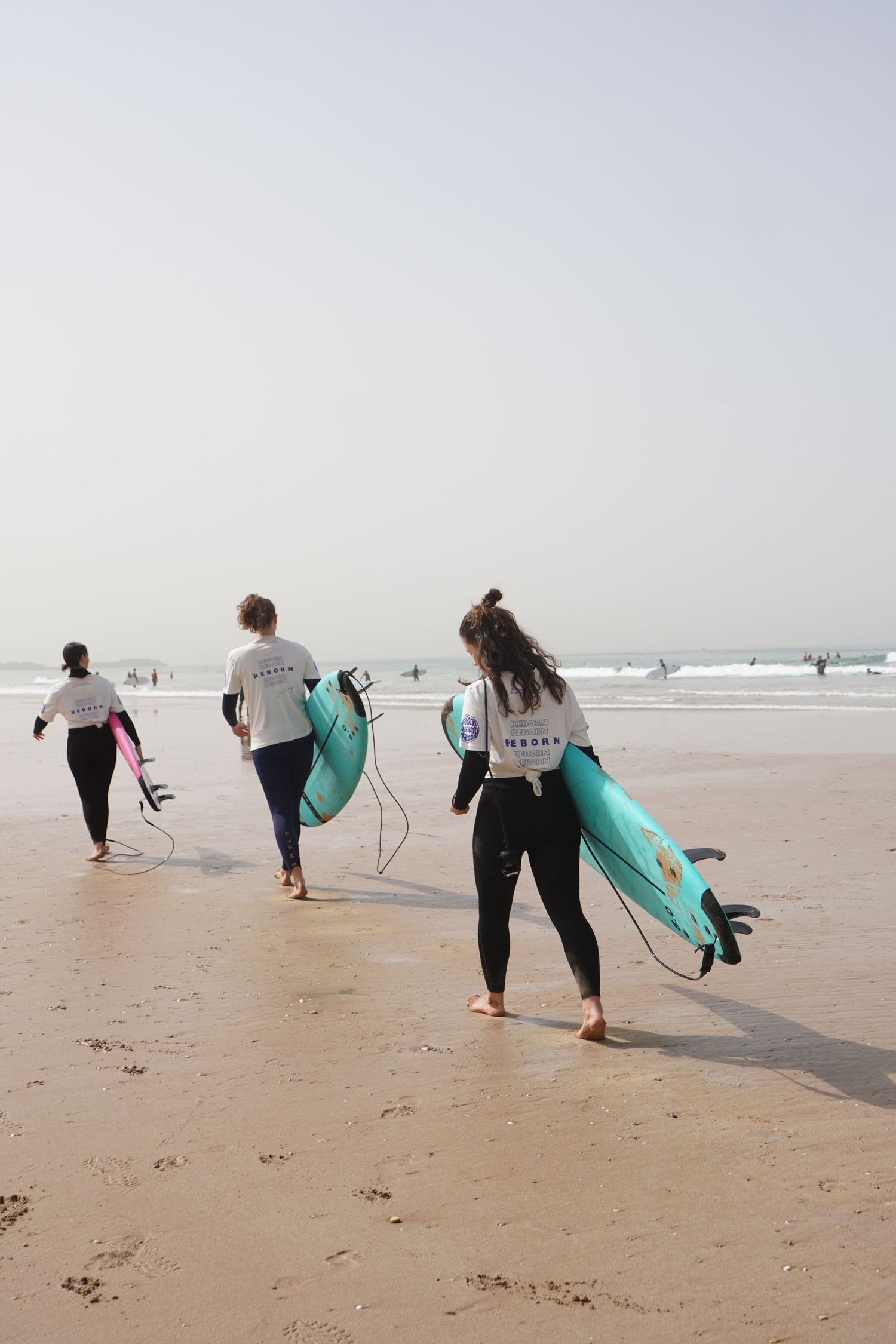 Three female surfers in wetsuits carrying colorful surfboards walking toward ocean waves on a sandy beach.