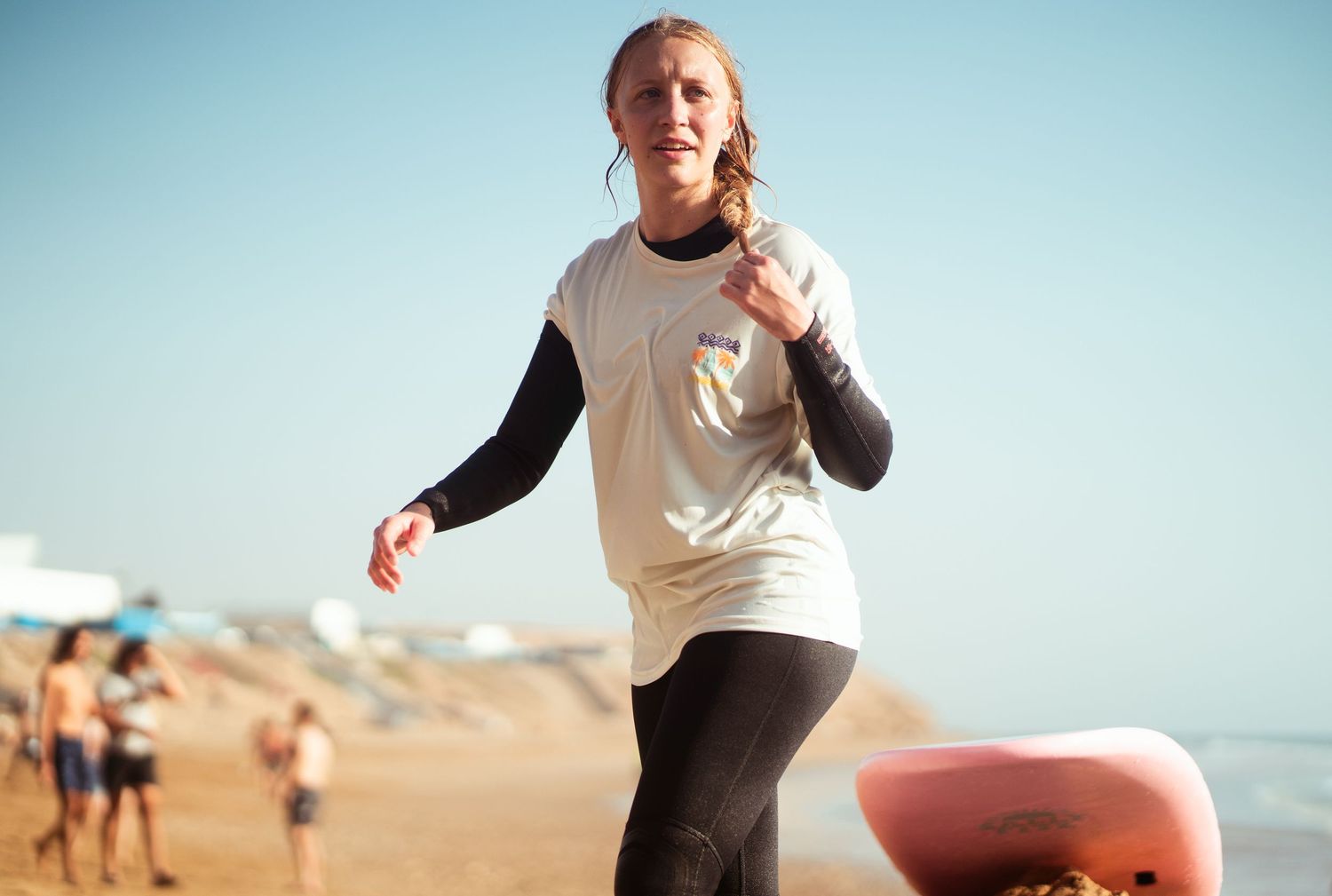 A female surfer in a white rash guard and black wetsuit runs along a sandy beach carrying a pink surfboard.