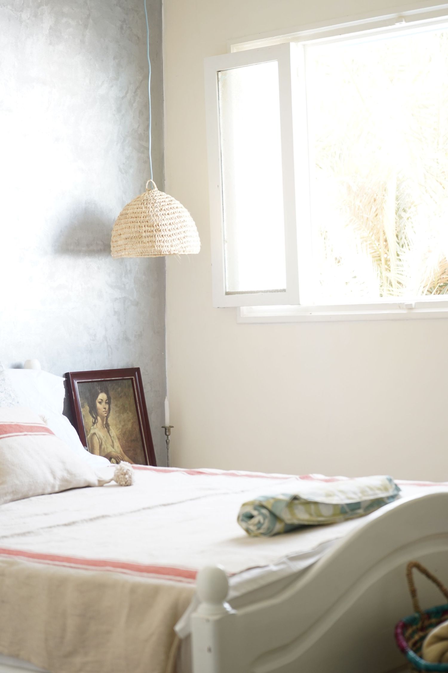 Bright, airy bedroom with white bedding, wicker pendant light, framed portrait, and natural light from a window.