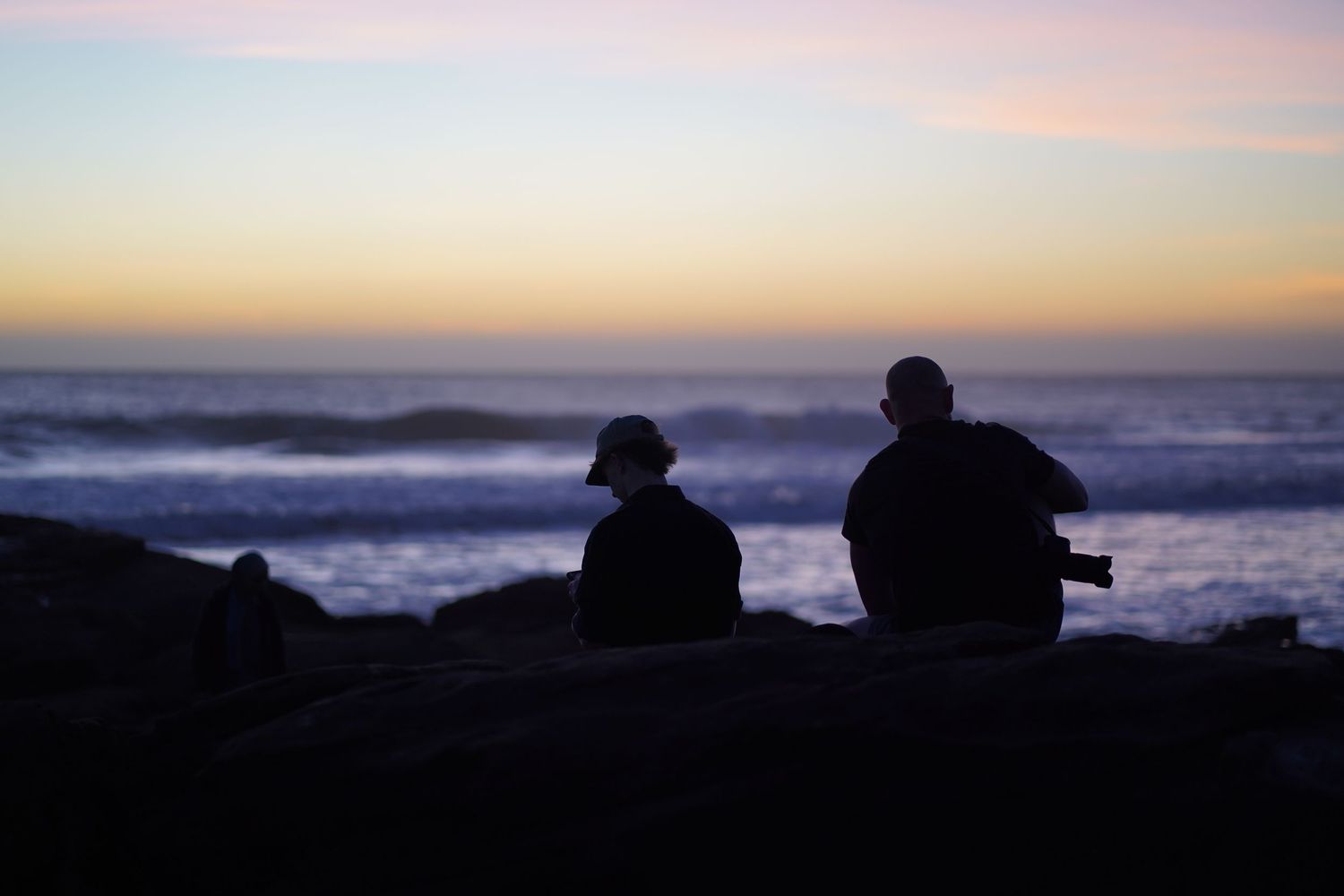 Two silhouetted figures sit on rocks watching a colorful ocean sunset with waves in the background.