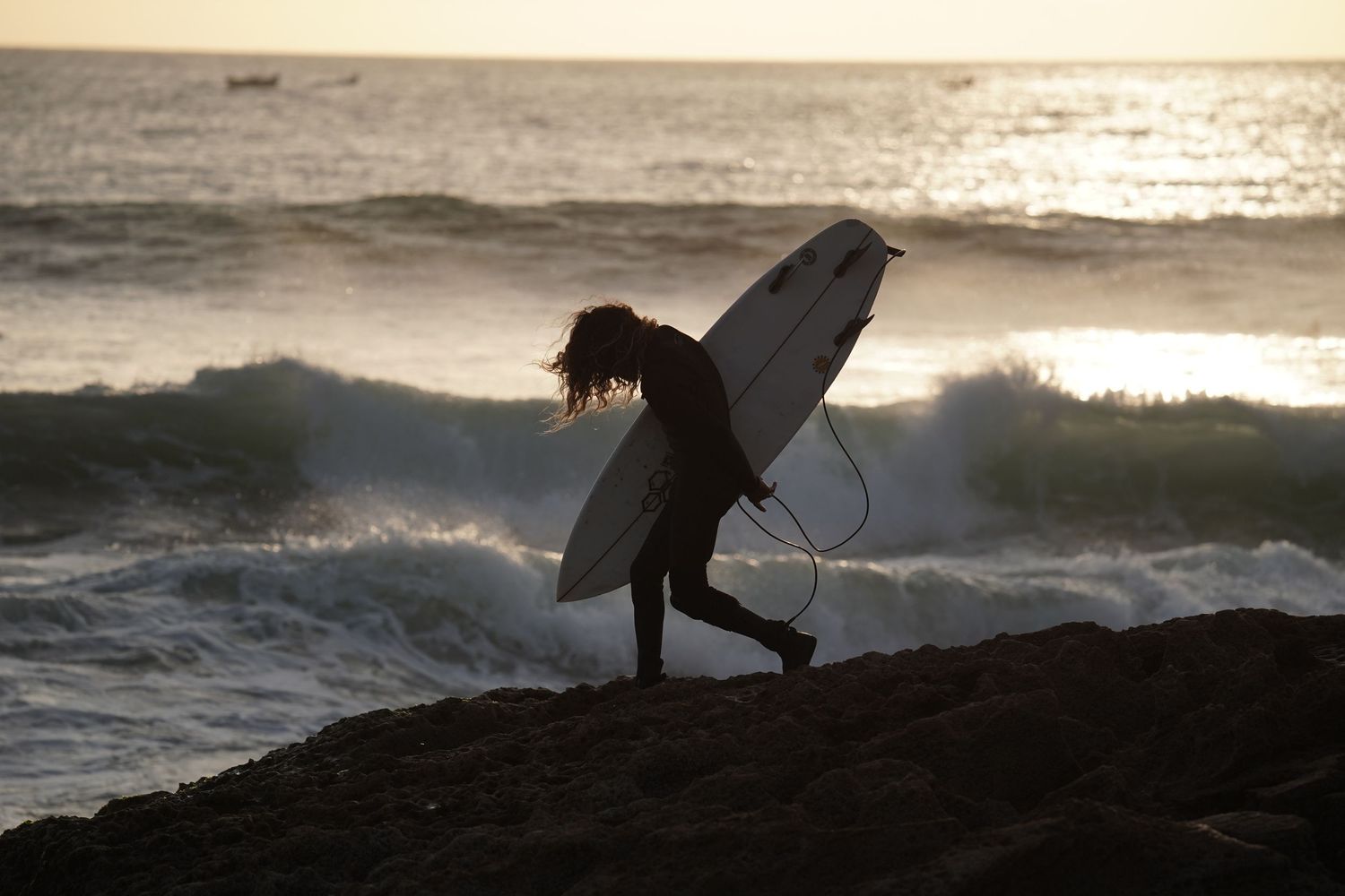 Surfer in wetsuit carrying a white surfboard across rocky shore with ocean waves at sunset.