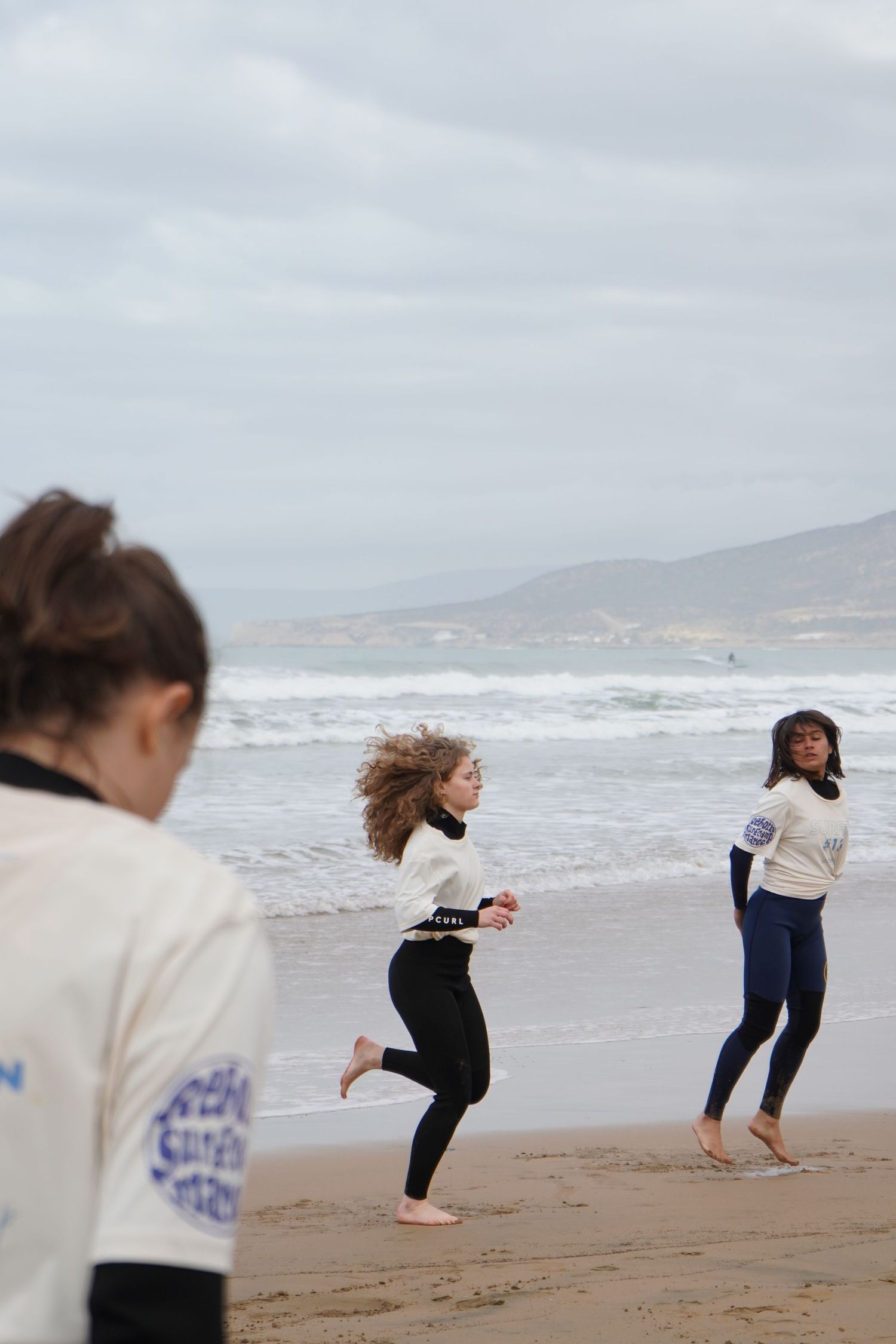 Two women in wetsuits running barefoot on a cloudy beach with ocean waves in the background.