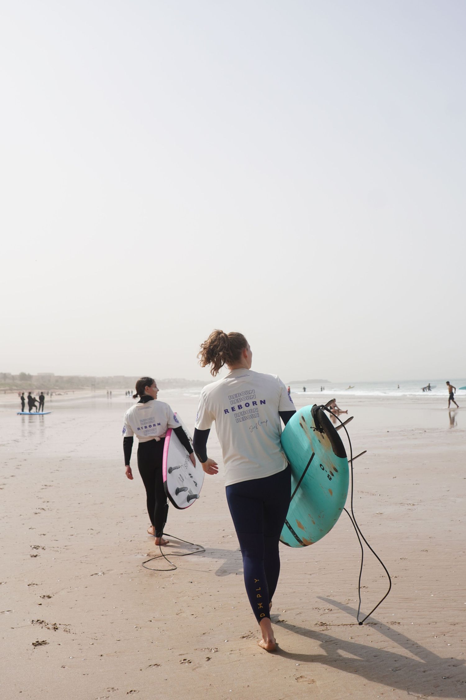 Two female surfers in white shirts carry colorful surfboards along a sandy beach toward ocean waves.