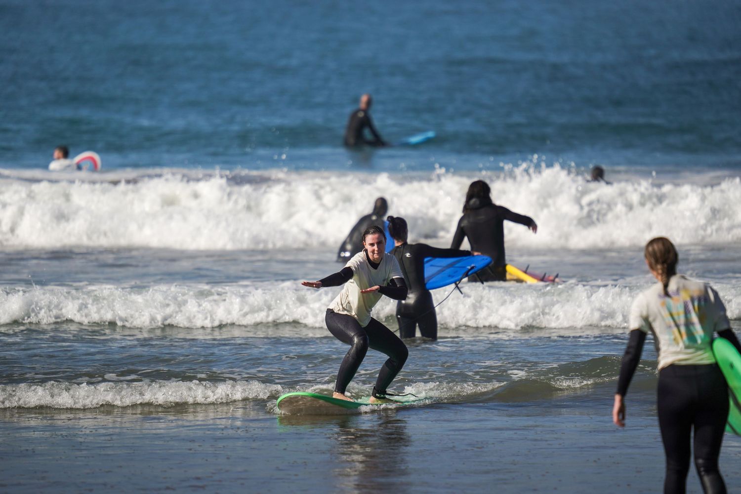 Surfers in wetsuits ride waves on a beach, with one person balancing on a surfboard while others carry boards nearby.