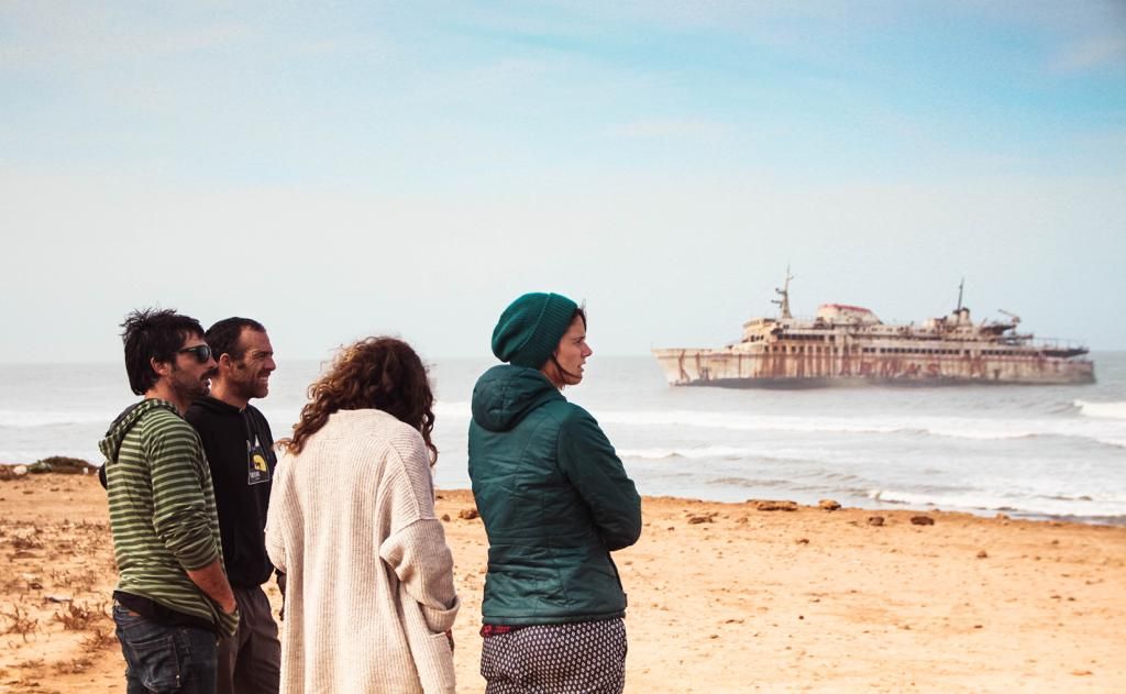 Four tourists stand on a sandy beach admiring a rusty shipwreck partially submerged in the ocean under a hazy sky.