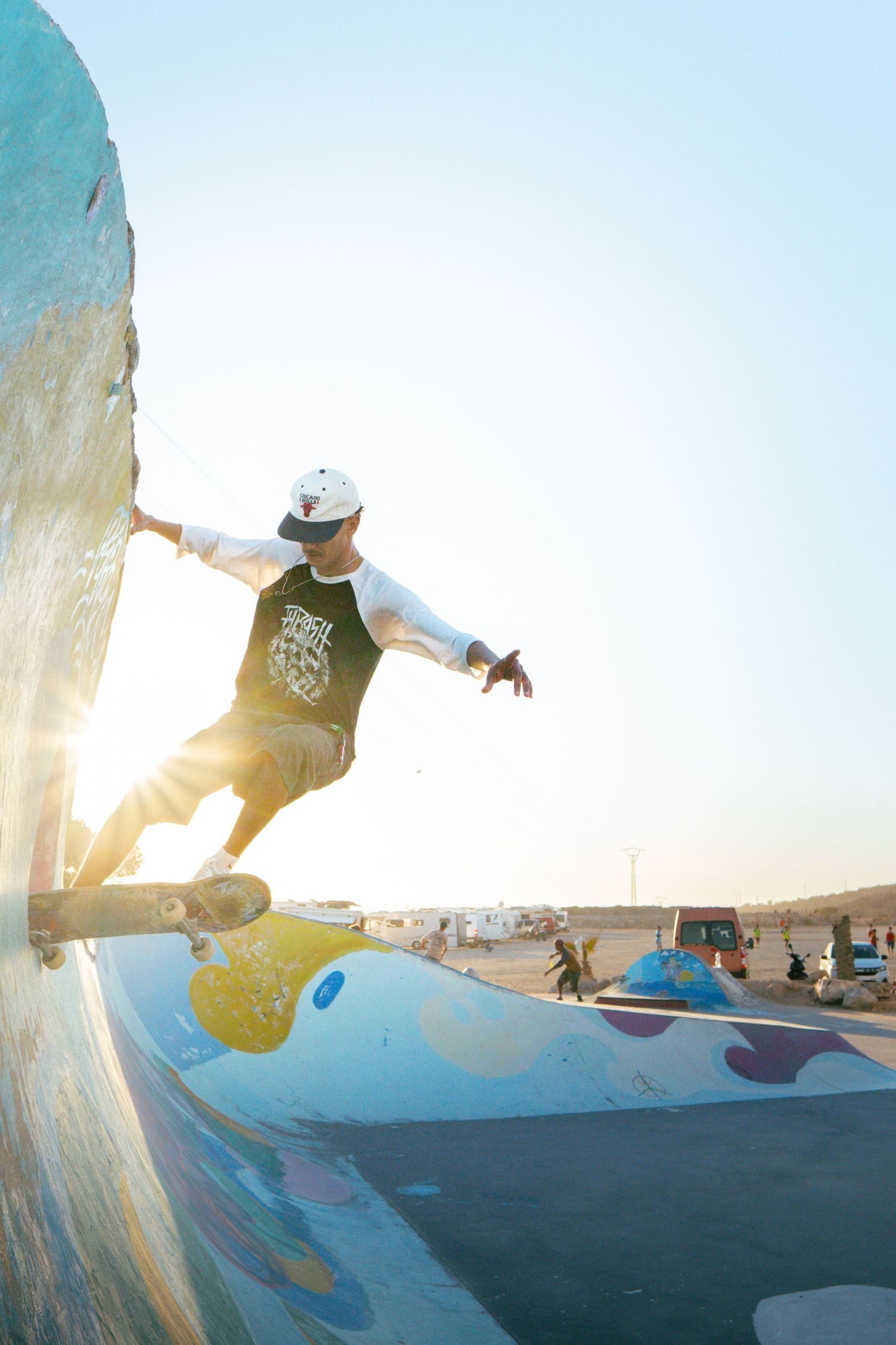 Skateboarder performing wall ride on colorful painted ramp at golden hour sunset outdoor skatepark.
