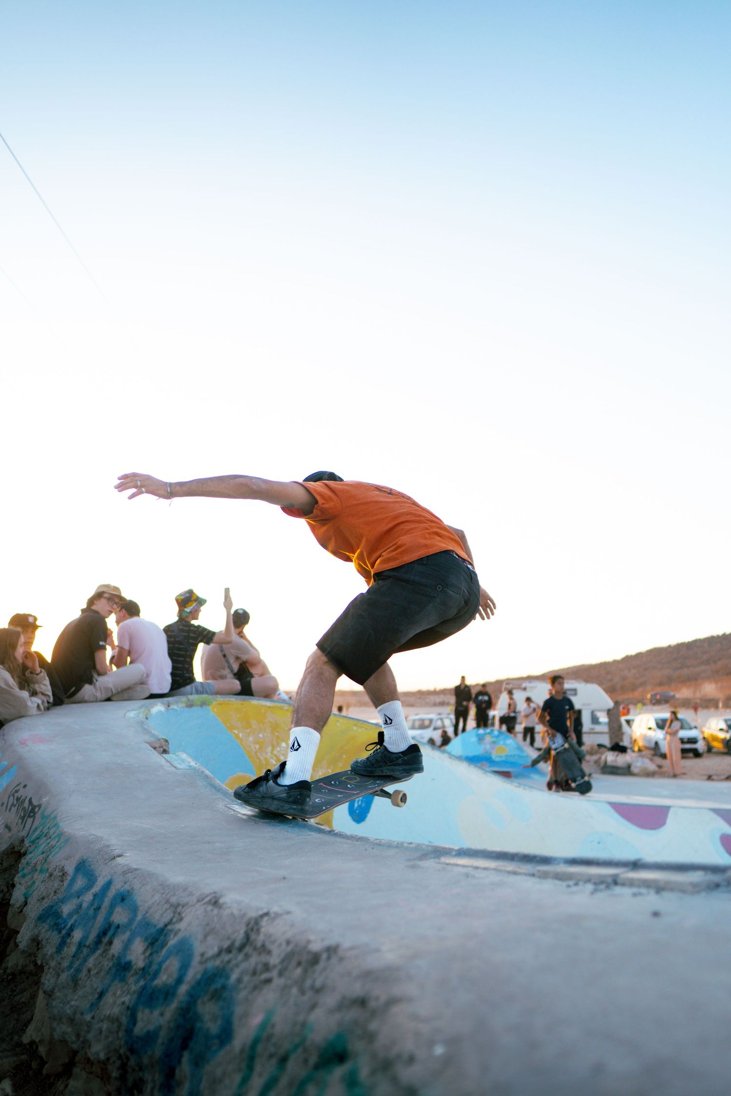 Skateboarder in orange shirt grinds on painted concrete ledge at outdoor skate park with spectators watching.