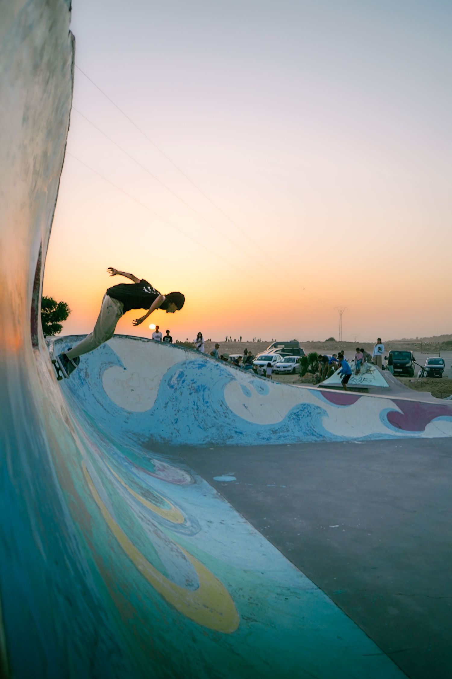 Skateboarder performing trick on painted wave mural wall at outdoor skate park during sunset.