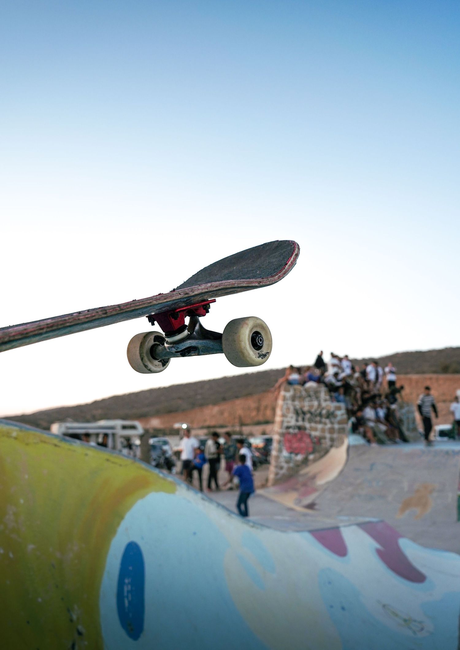 Close-up of a skateboard deck and wheels airborne above a colorful ramp with crowd visible in background.
