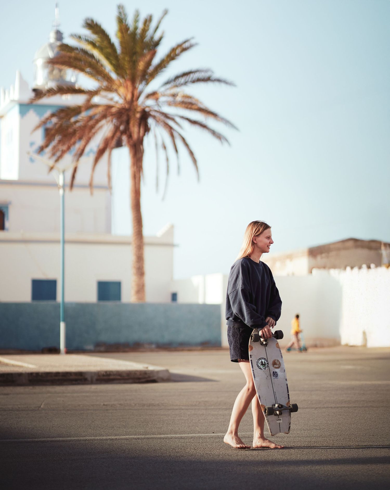 A barefoot blonde woman holds a longboard skateboard on a sunny street with a palm tree and white buildings.