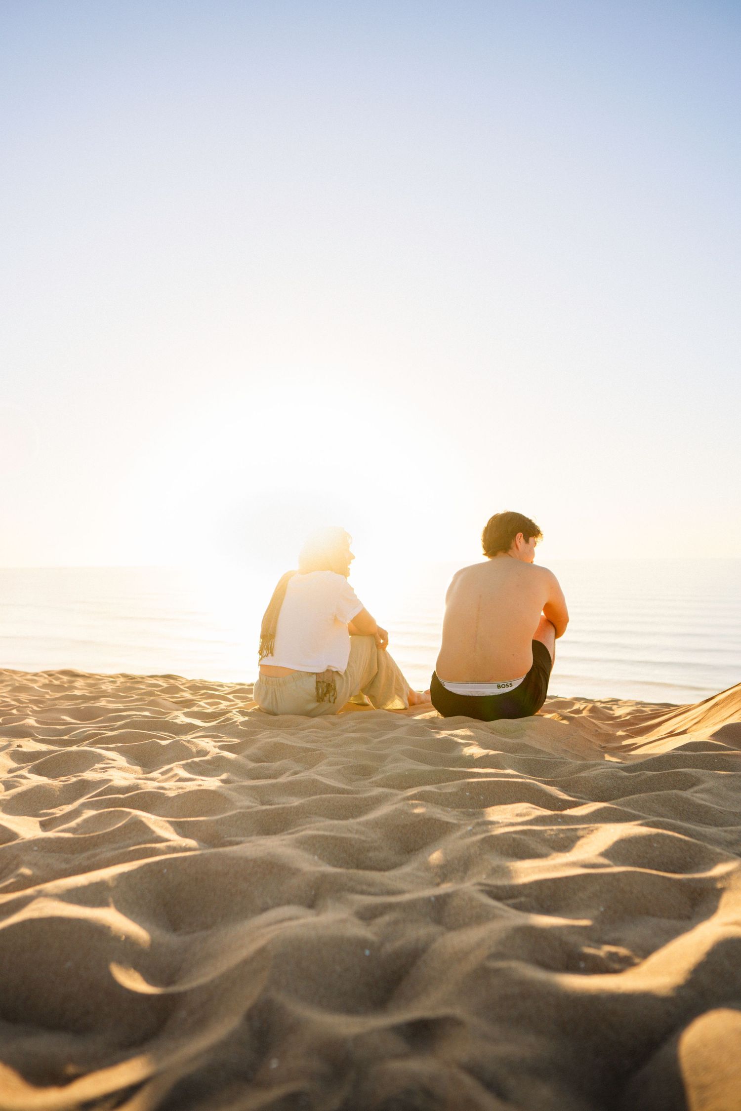 Two people sitting on sandy beach at golden sunset overlooking calm ocean waters.