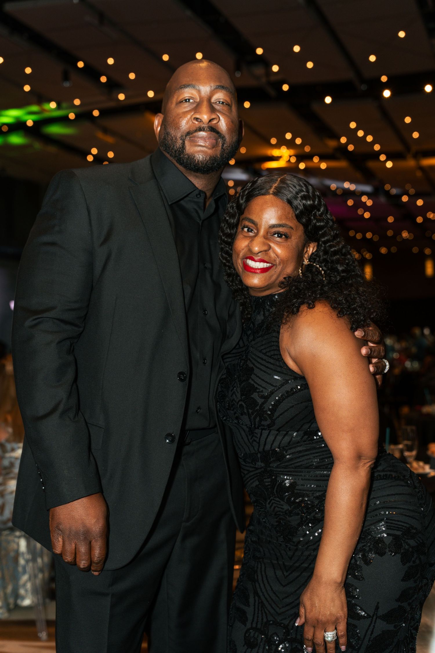 Couple dressed in matching black formal attire smile together at an elegant evening gala event.