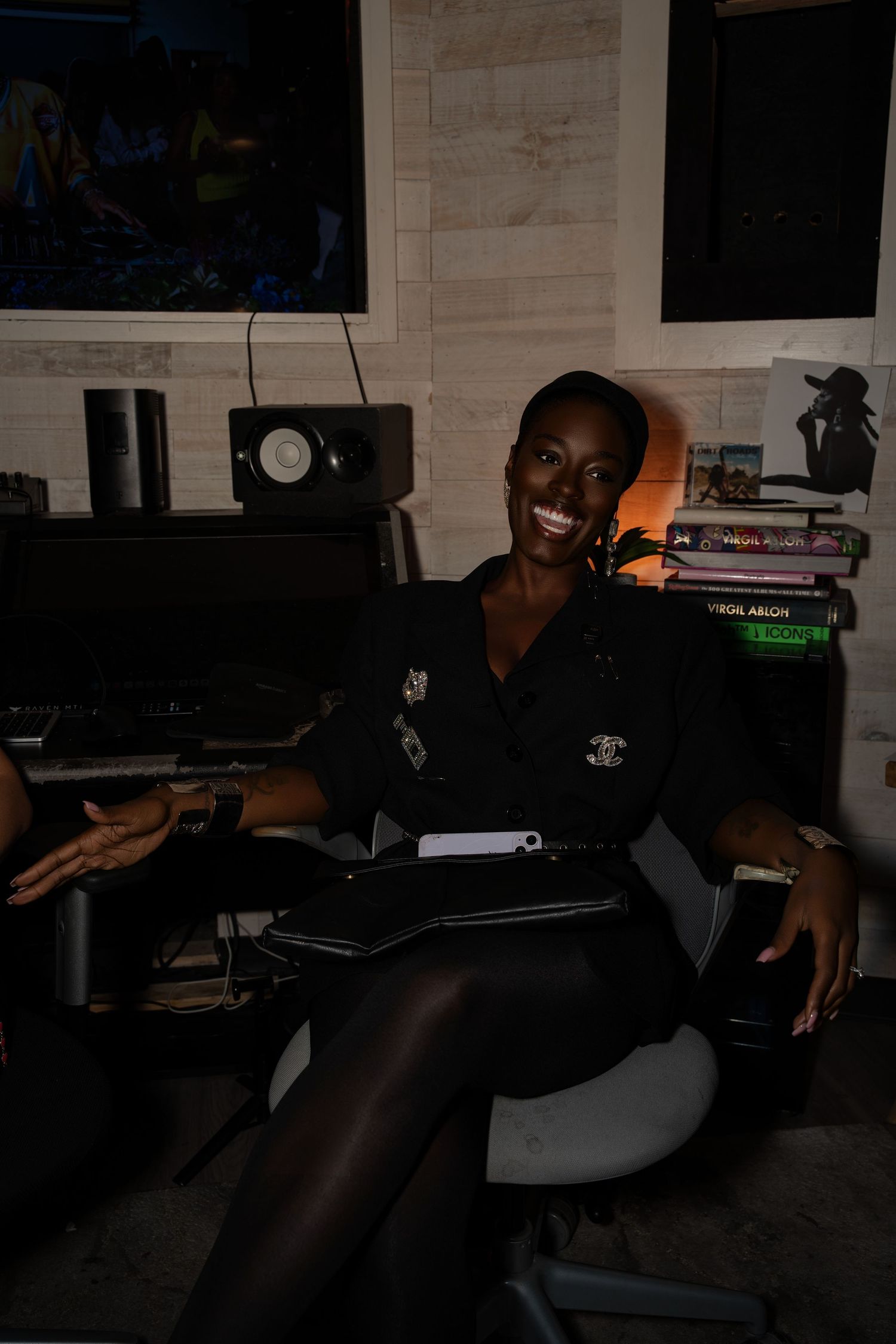 Smiling woman in black outfit sitting at a desk in a dimly lit studio room with monitors and equipment.