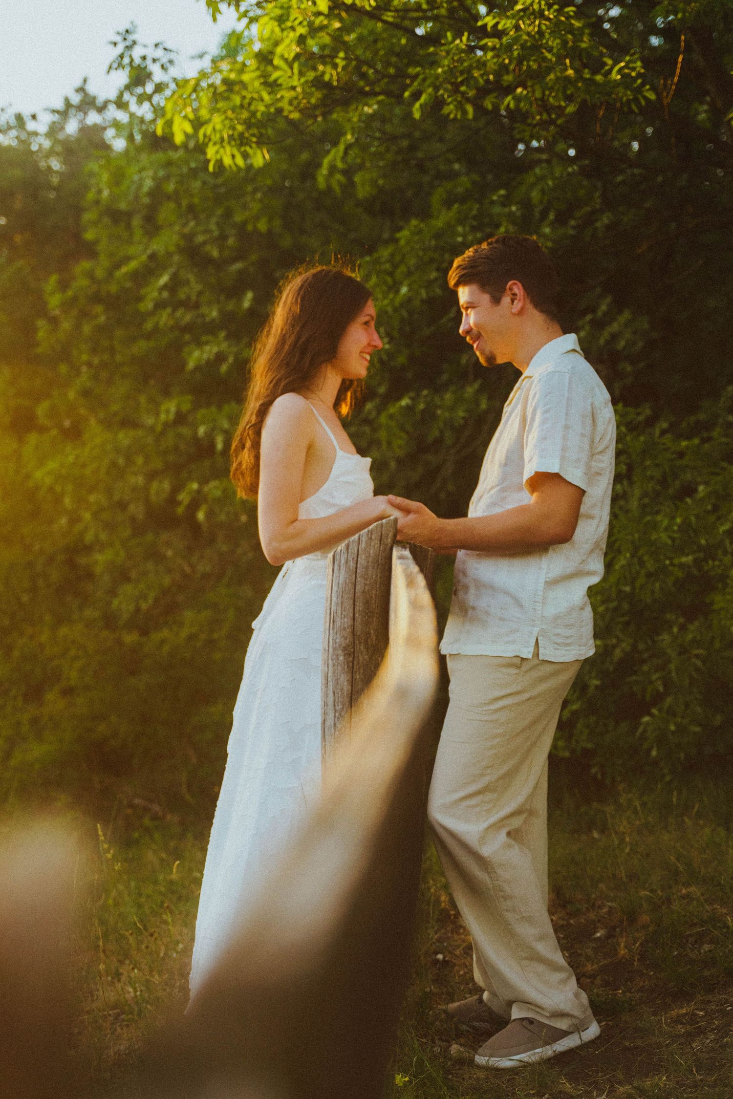 Couple smiling and holding hands in golden sunset light surrounded by lush green garden foliage.