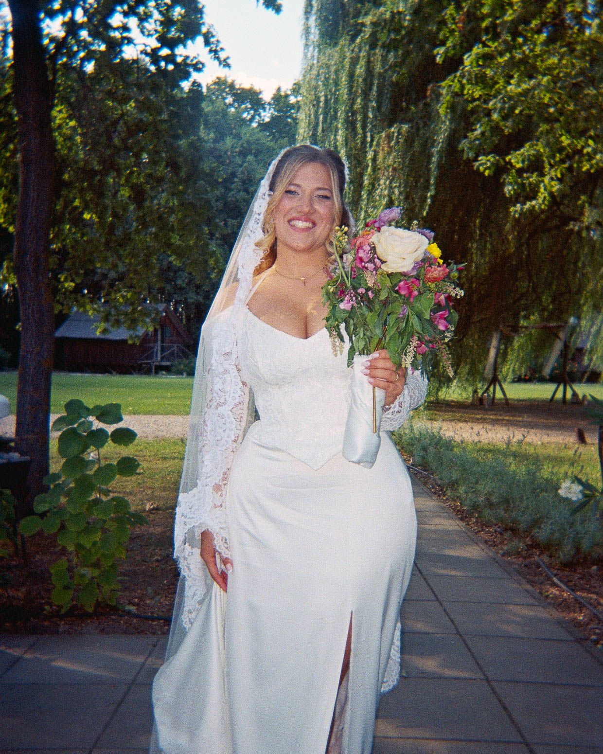 Smiling bride in white lace wedding dress and veil holding colorful bouquet outdoors among green trees.