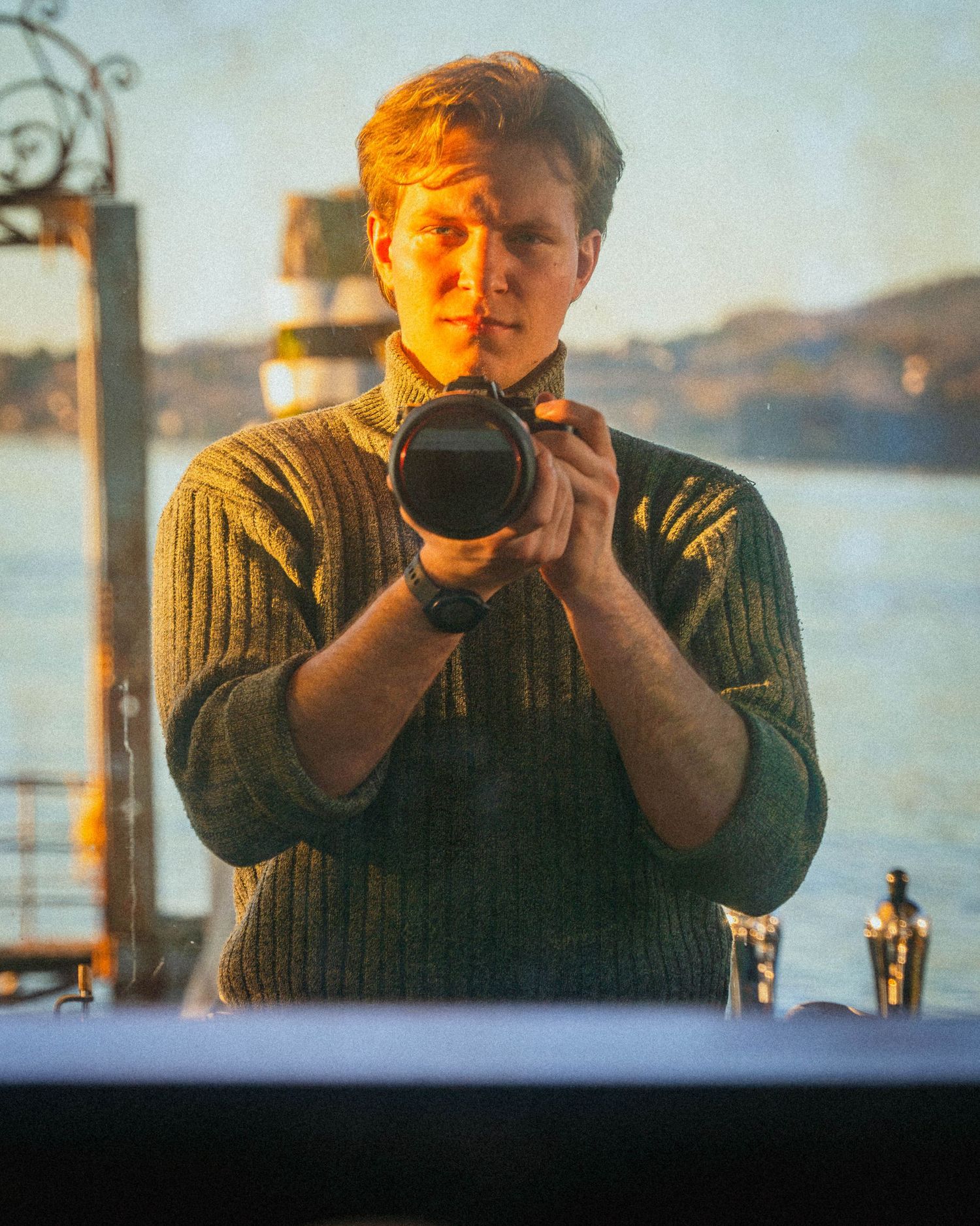 Photographer taking a mirror selfie with a camera during golden hour by the waterfront.