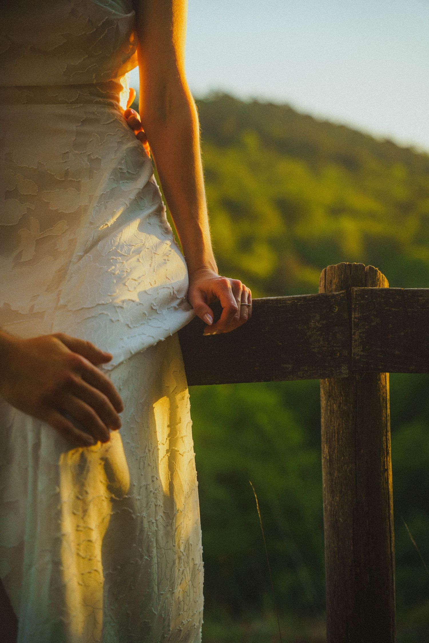 Pregnant woman in white dress holding wooden fence at sunset.