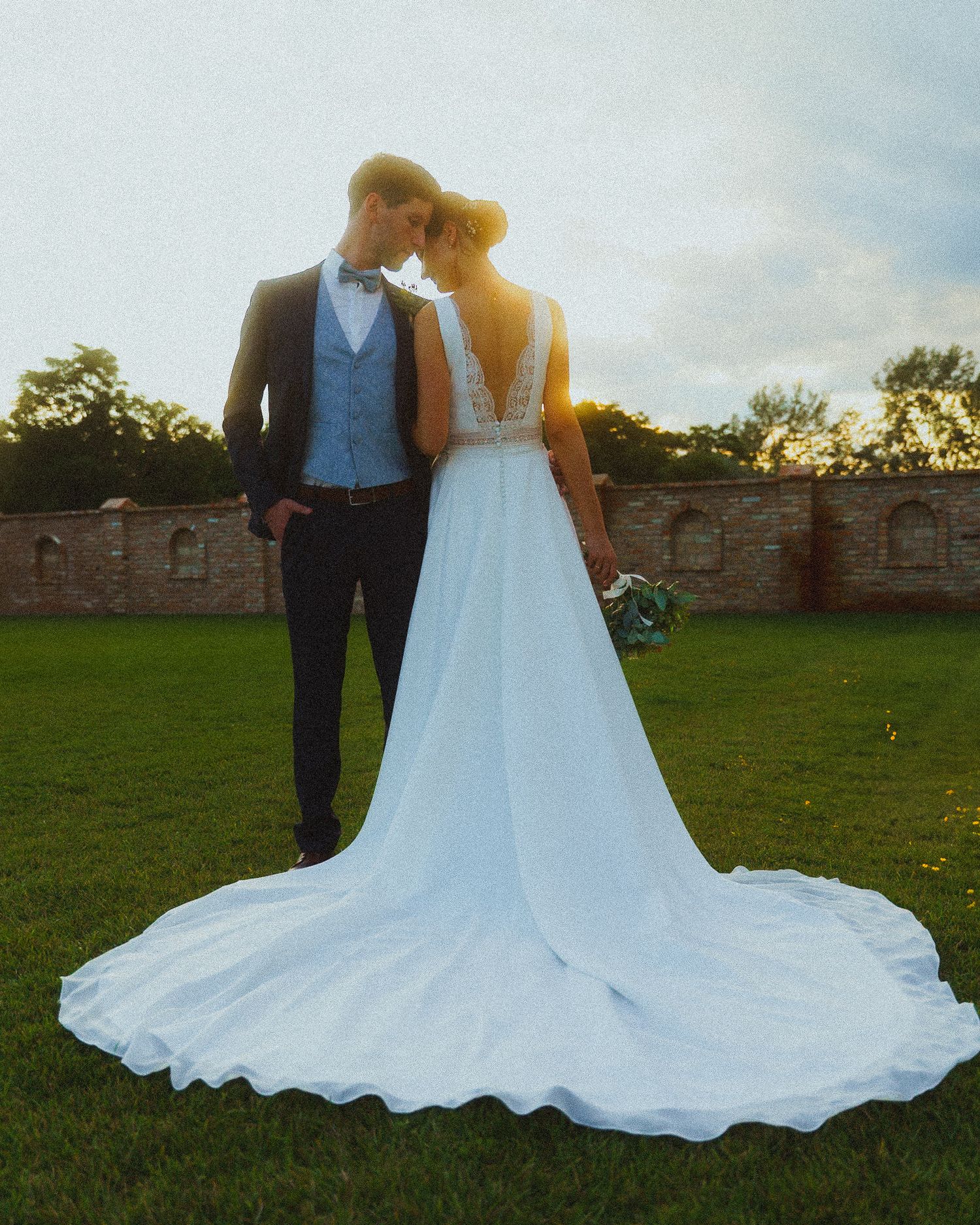 Bride in flowing gown with long train stands with groom at magical golden hour wedding.