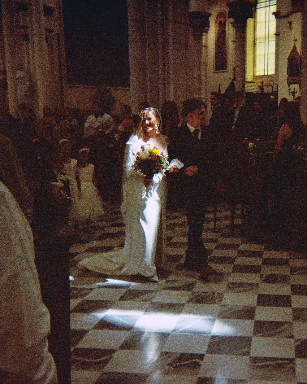 Bride in white gown walks down church aisle with groom, guests seated in pews, checkered marble floor.