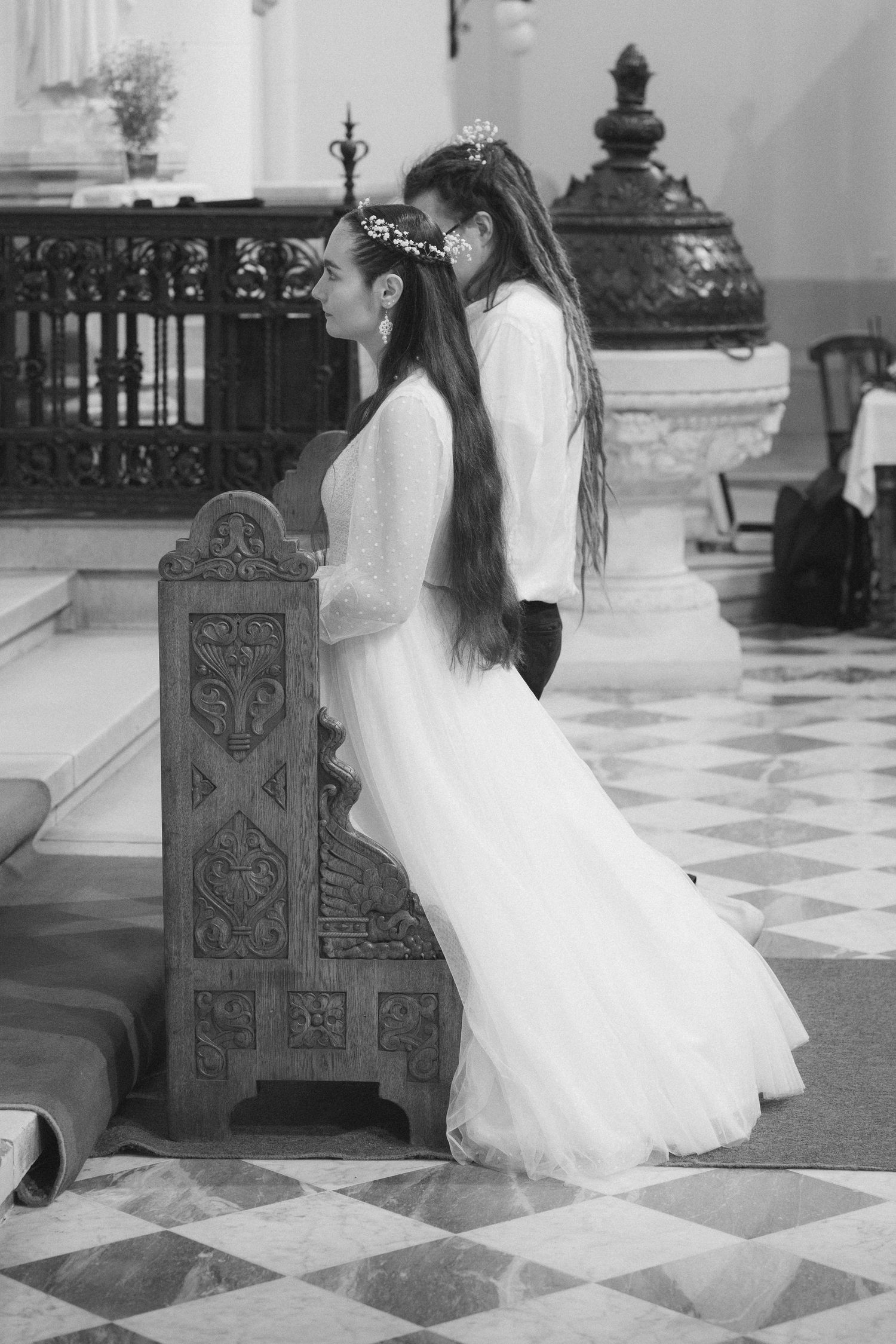 Bride with floral crown kneeling in prayer at a church pew, black and white wedding photo.