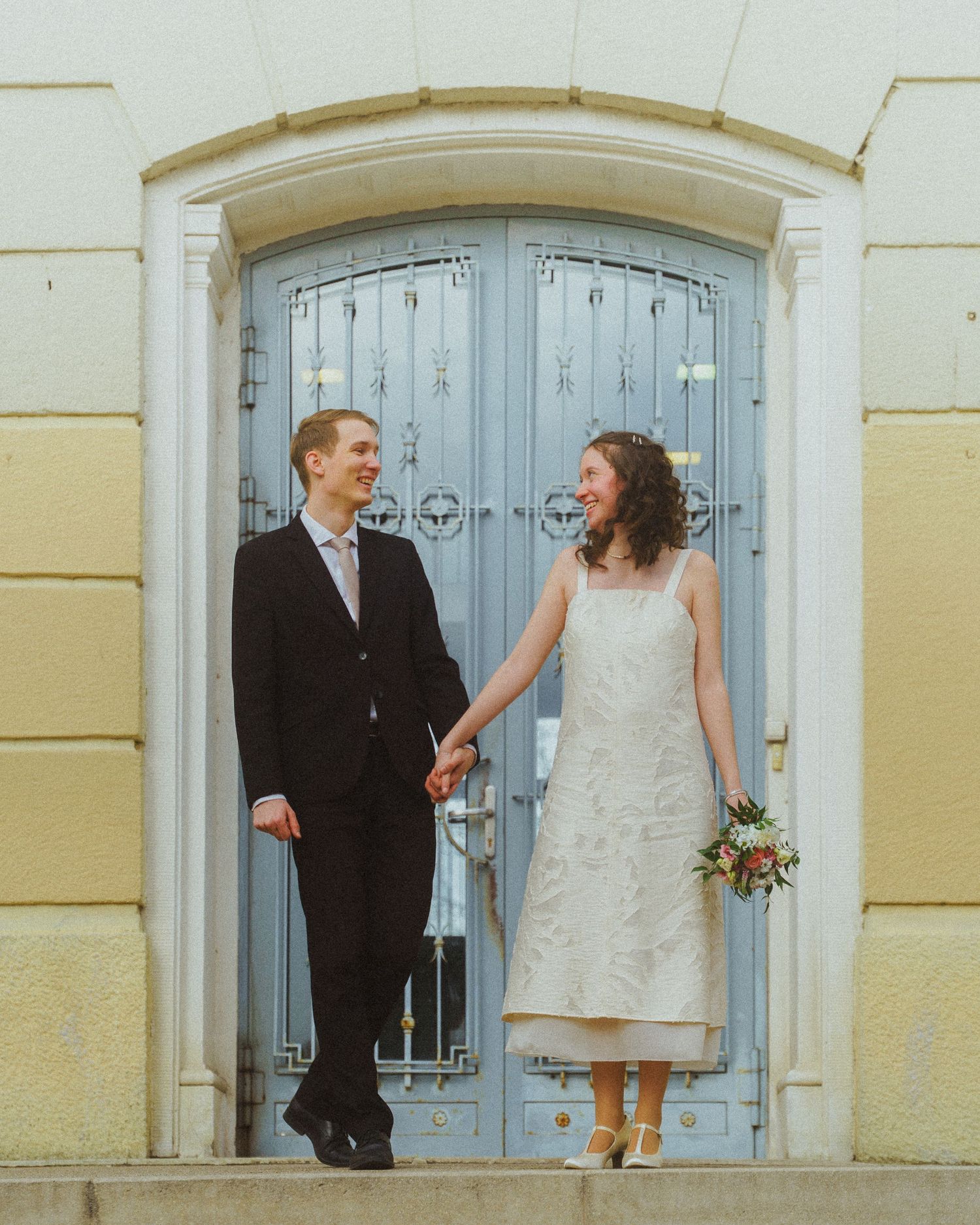 Happy newlywed couple holding hands with bride holding bouquet in front of a blue ornate door.