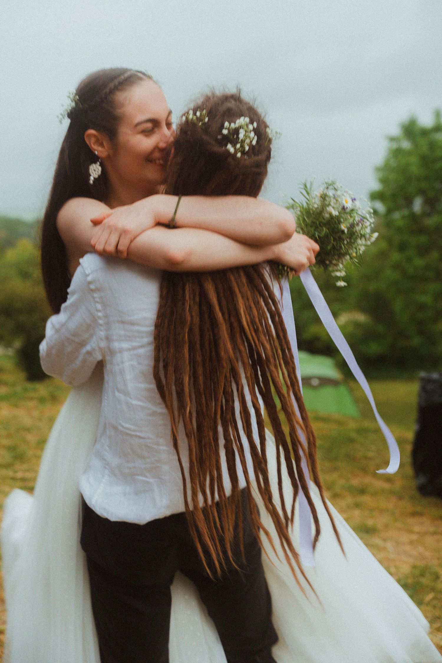 Two women embracing warmly outdoors, one in a white wedding dress holding a floral bouquet.
