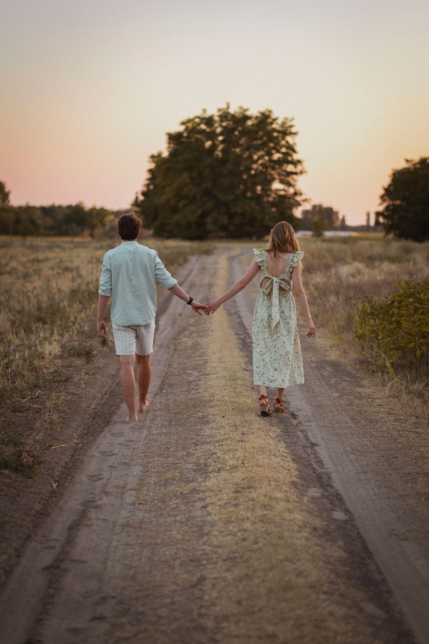 Couple holding hands walking down a rural dirt road at sunset, viewed from behind.