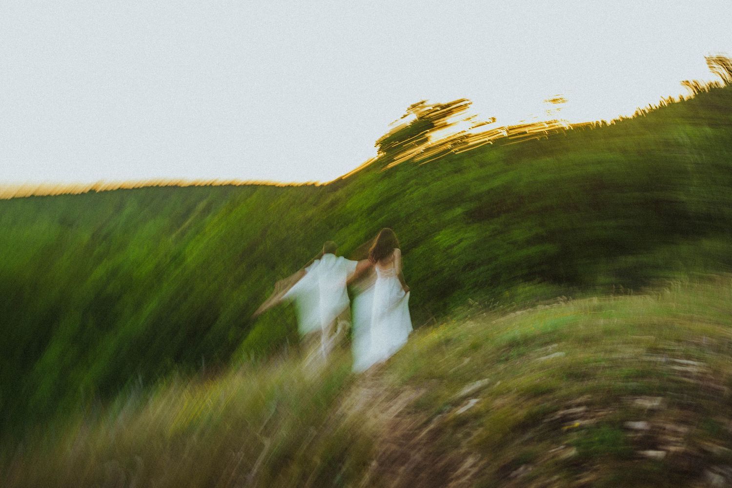 Two women in white dresses running through a green hillside field captured with motion blur effect.