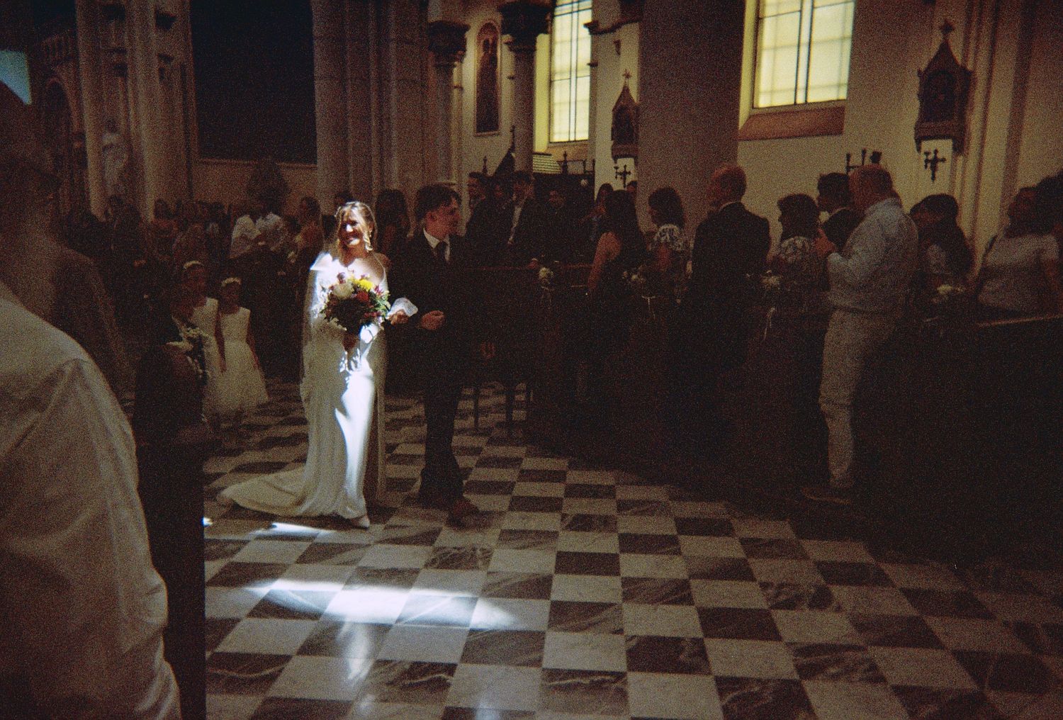 Bride and groom walk down the aisle of a historic church with checkered floors during their wedding ceremony.
