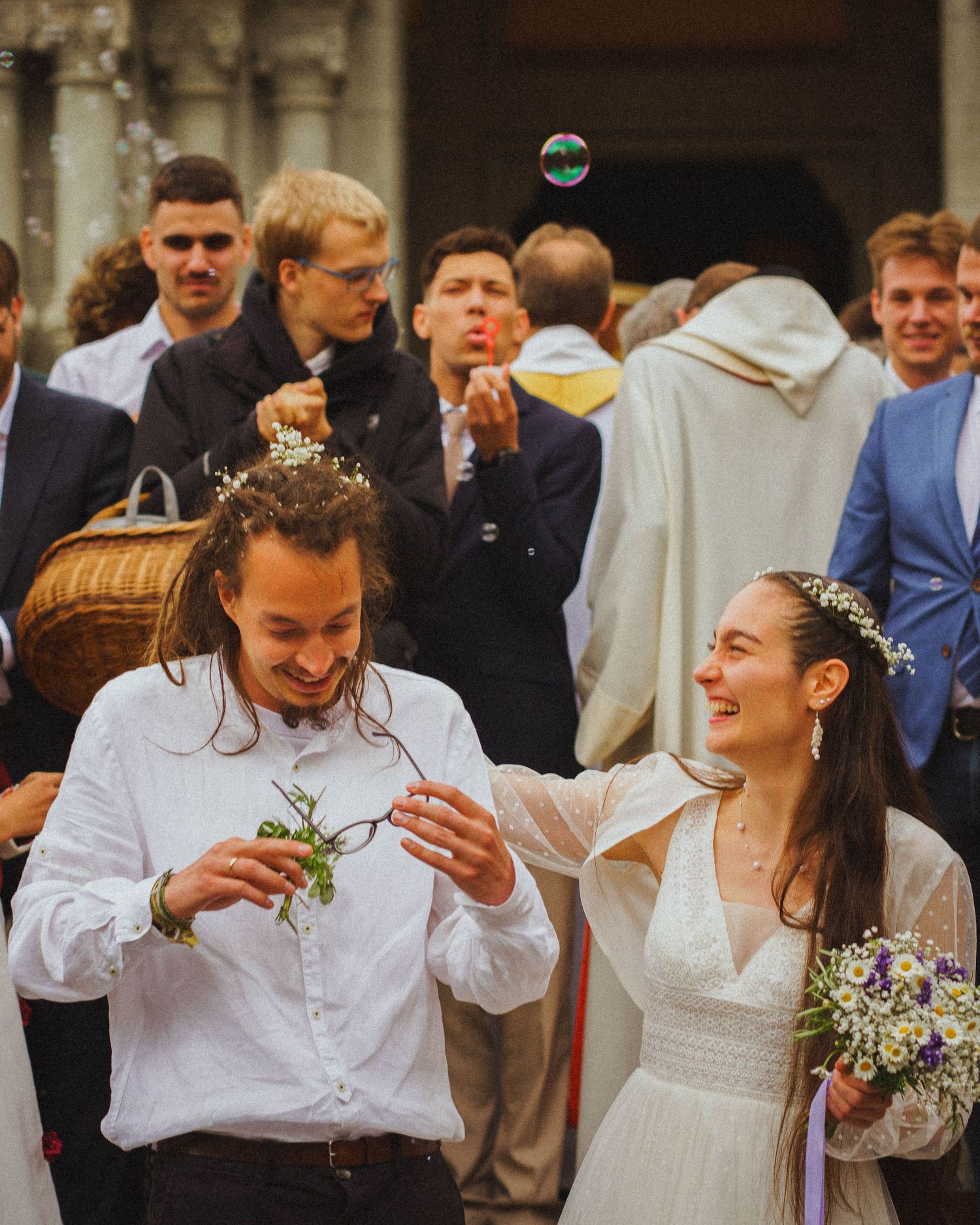 Smiling bride in white dress with flower crown and groom celebrating outside church with bubbles.