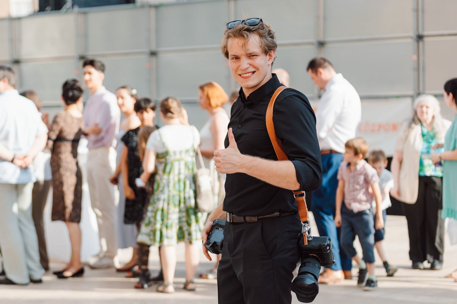 Young male photographer smiling with cameras at an outdoor event on a sunny day.