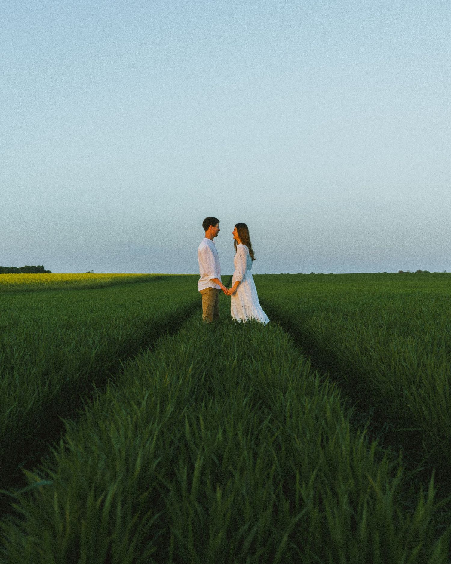 Couple holding hands facing each other in a lush green field at twilight.
