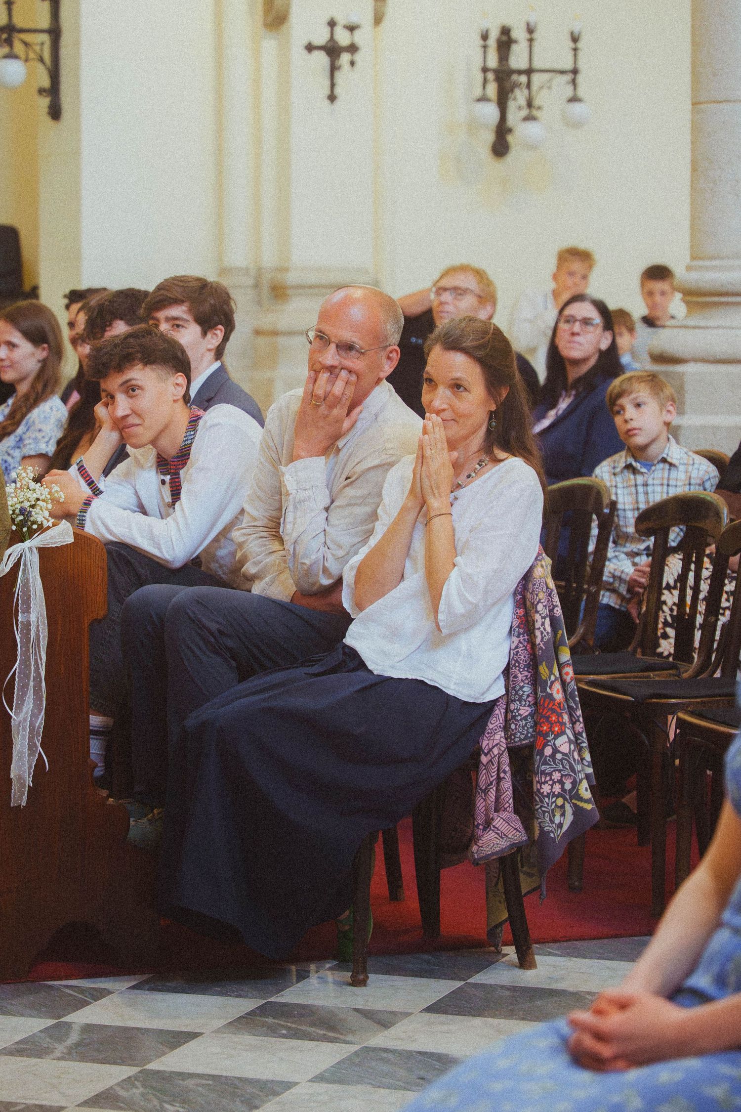 Wedding guests seated in a church pew, smiling and watching the ceremony with hands clasped in anticipation.