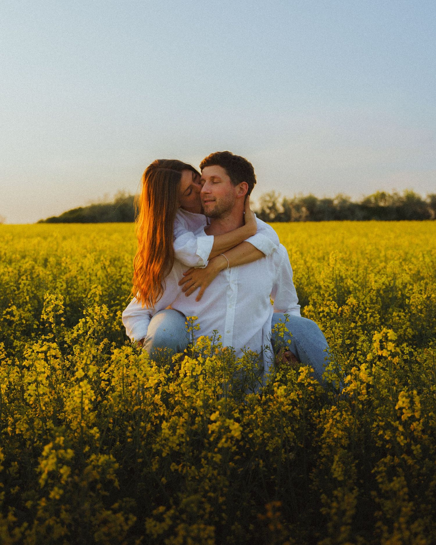 Woman hugging man from behind while sitting together in a sunny yellow rapeseed field.