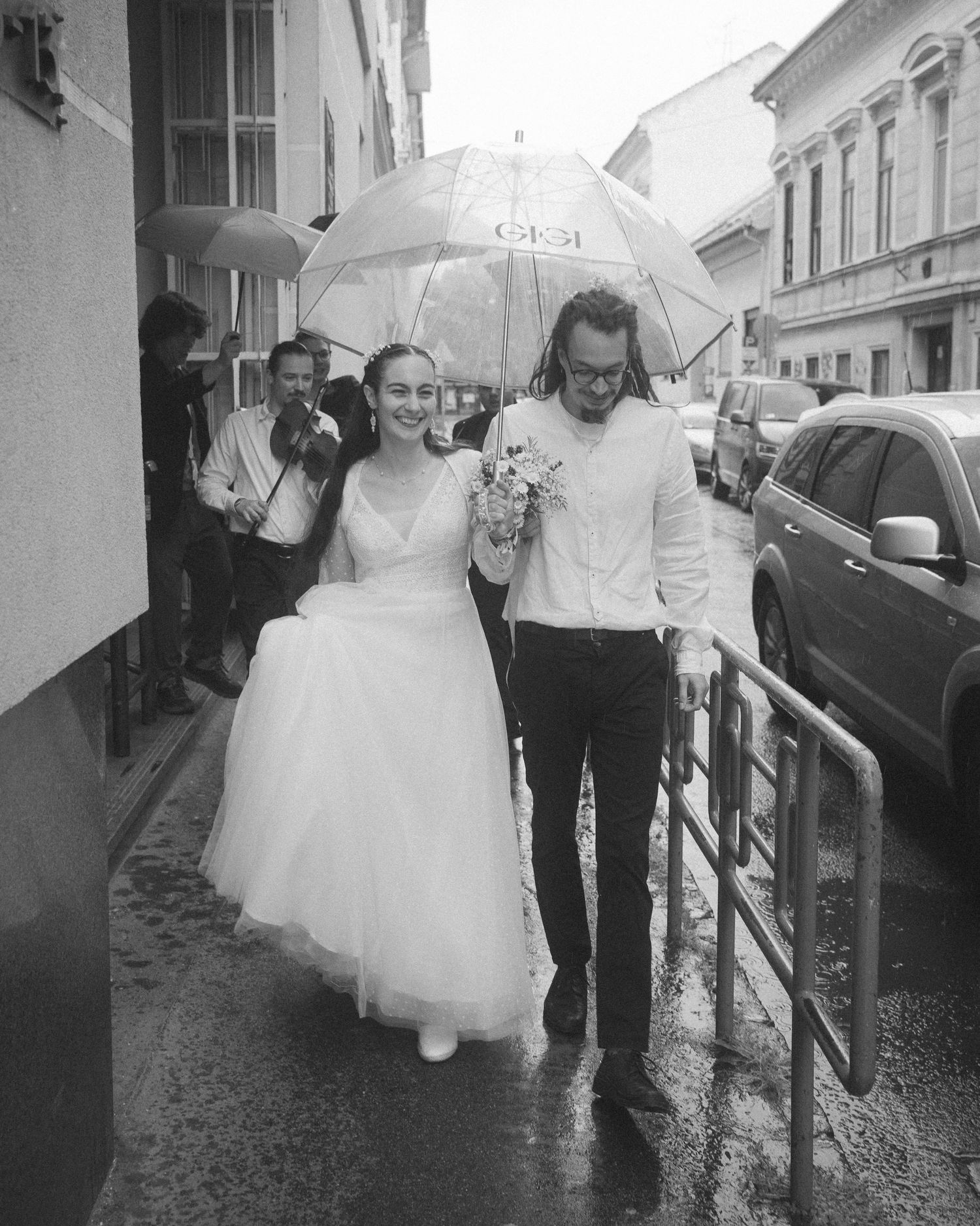 Black and white photo of a bride and groom walking in the rain under a clear umbrella on a wet city street.