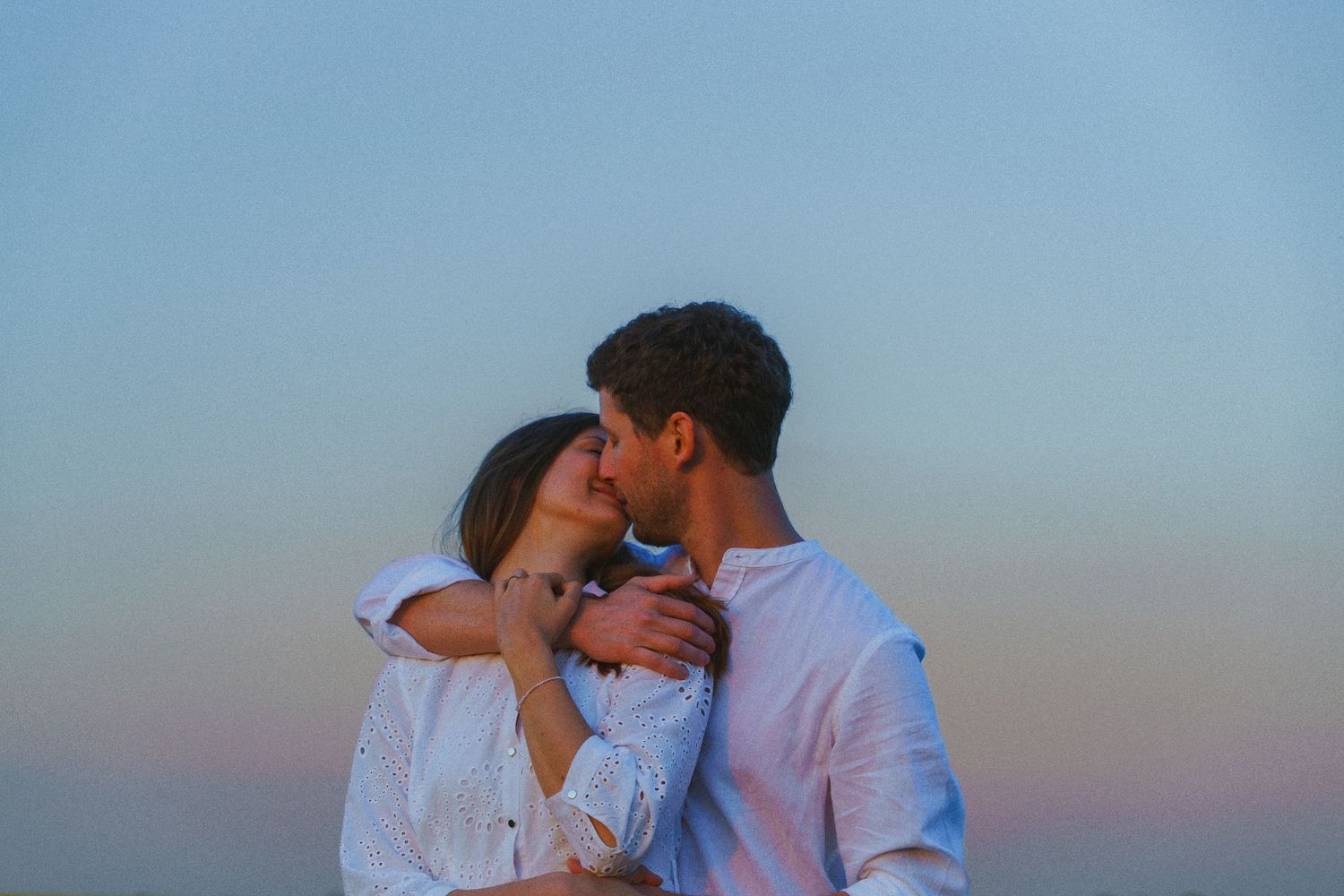 Romantic couple embracing and almost kissing against a soft pastel dusk sky.