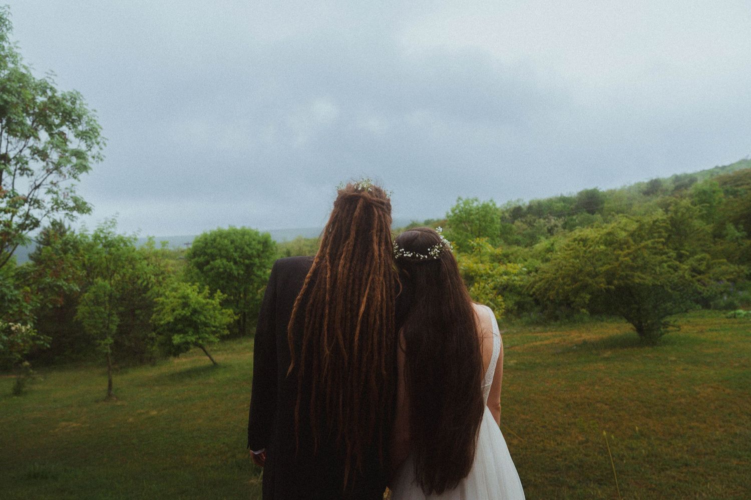 Two women with long hair wearing flower crowns standing in a green meadow, viewed from behind.