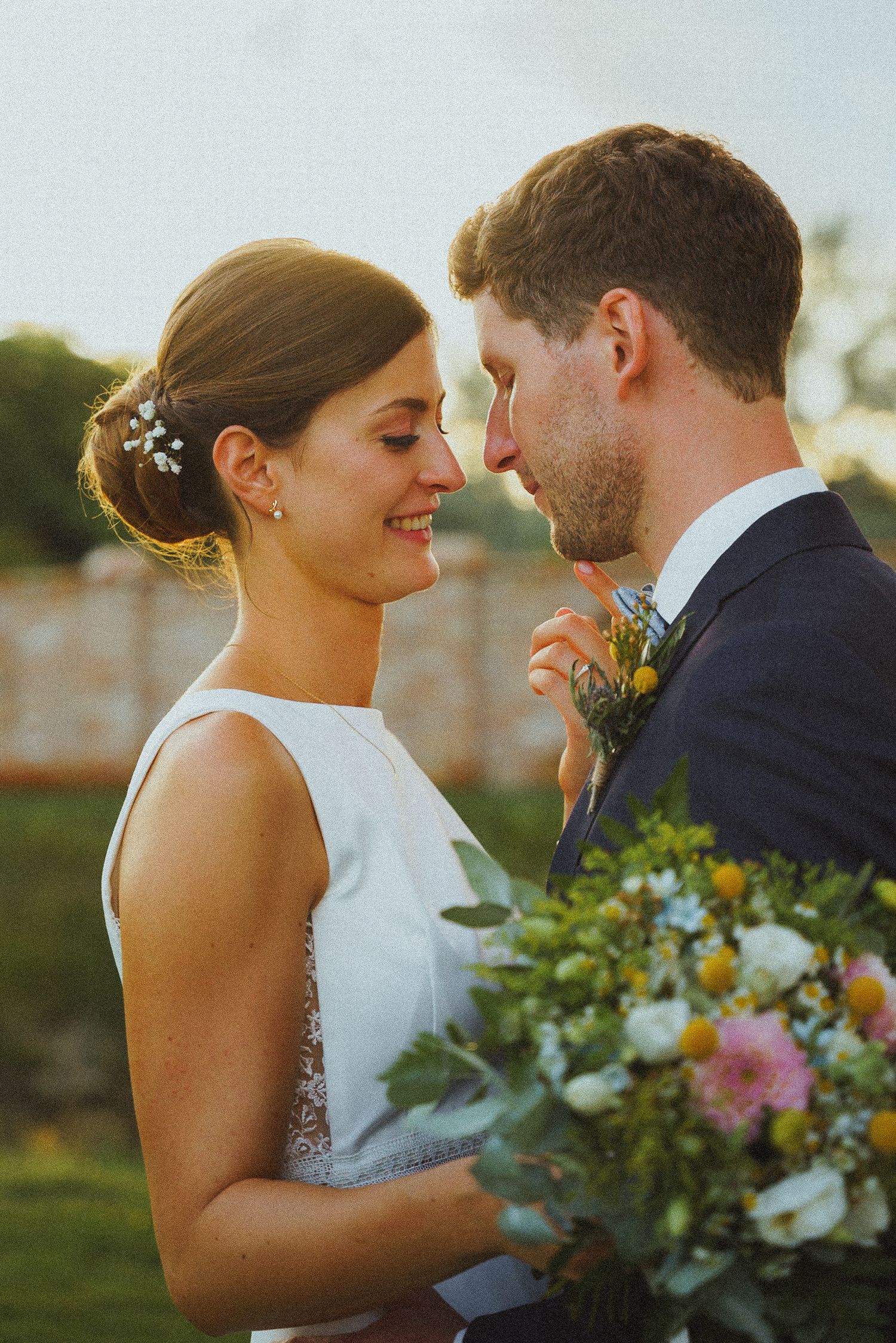 Smiling bride holds colorful bouquet while sharing tender moment with groom at sunset wedding.