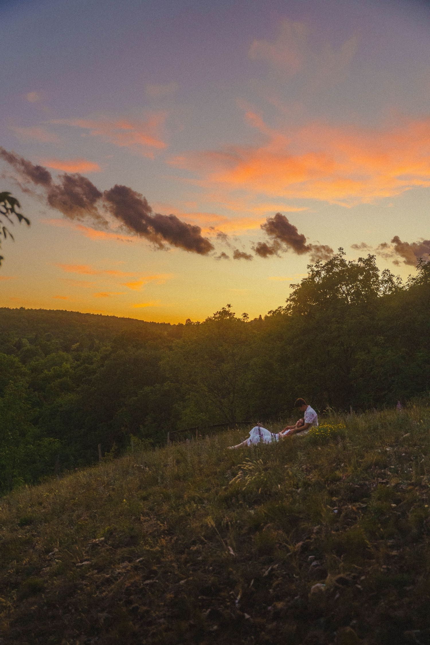 Couple lying on hillside under vibrant orange and purple sunset sky.