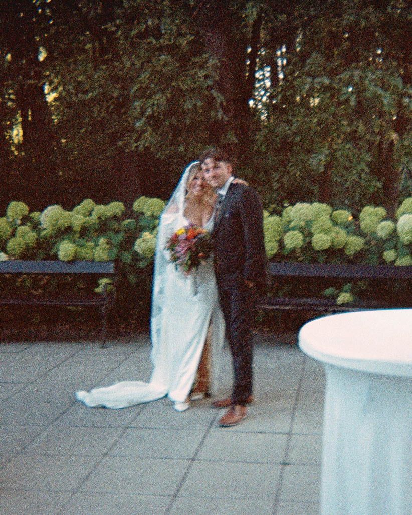 A bride and groom pose together outdoors at dusk, surrounded by lush hydrangea bushes on their wedding day.
