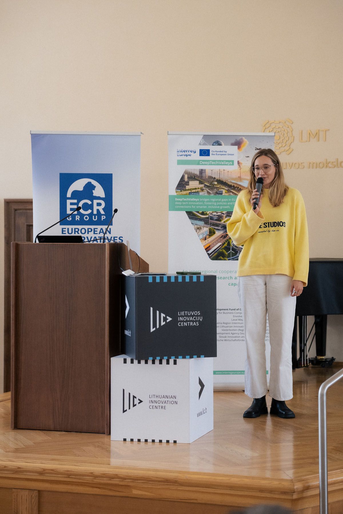 Woman in a yellow sweater and white pants presents on stage next to ECR Group and Lithuanian Innovation Centre displays.