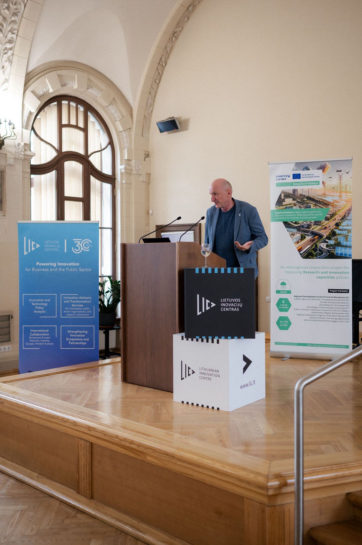 Bald man in a blazer speaks at a wooden podium on stage next to Latvian Innovation Centre branded boxes.
