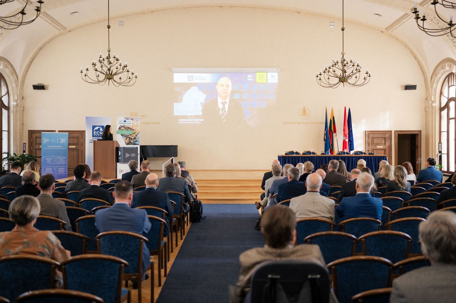 Audience seated in blue chairs watches a presenter on a large screen in a chandelier-lit conference hall.