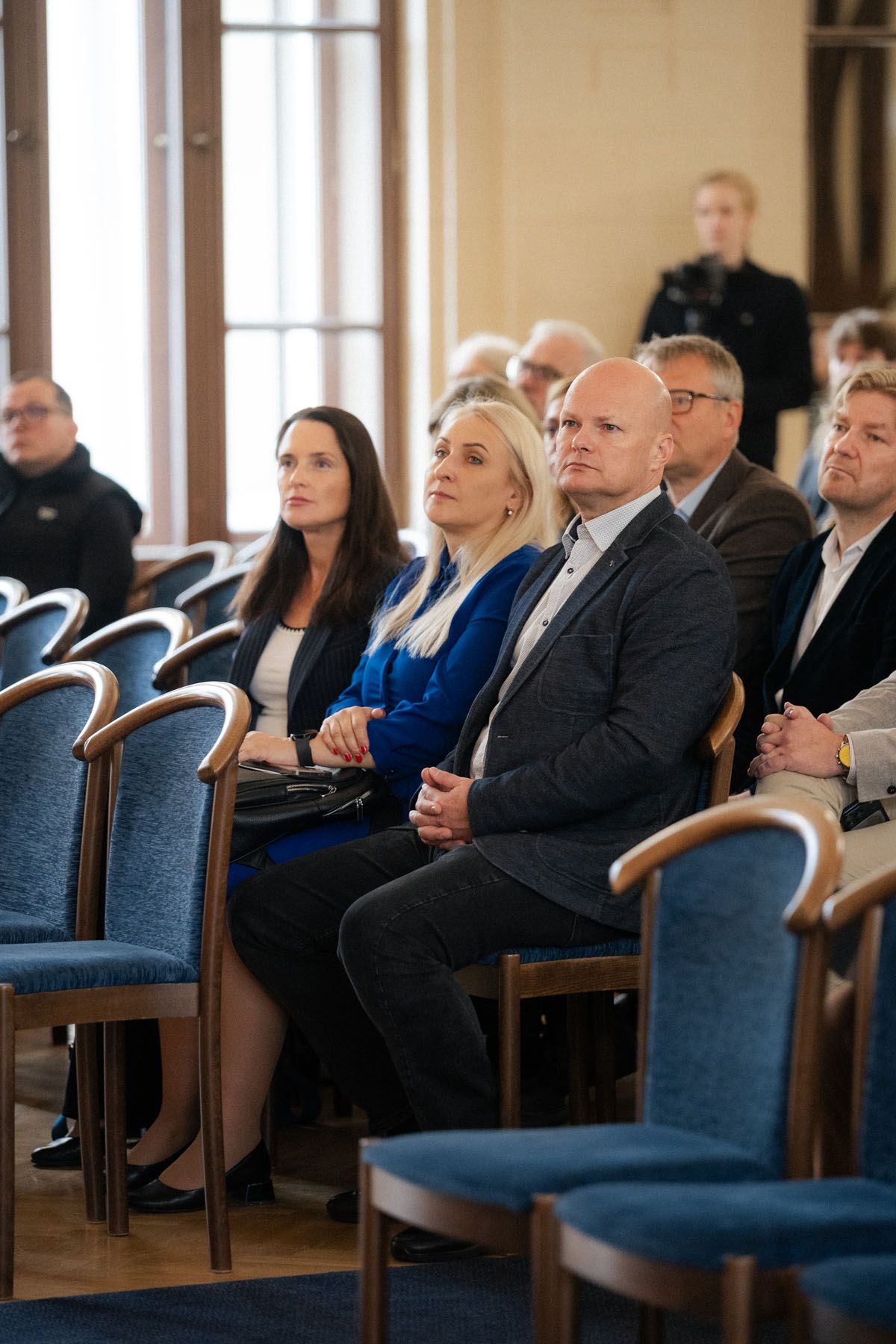 Attendees sit in blue chairs in a bright conference hall, including a bald man in a gray suit and two women beside him.