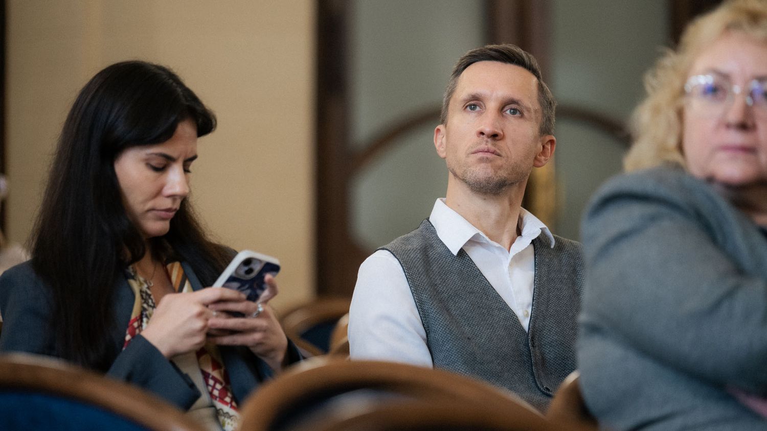 Audience members at a conference, including a woman checking her phone and a man in a vest looking toward the stage.