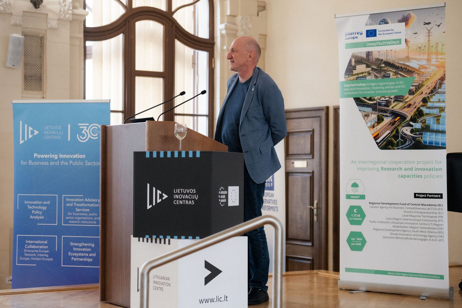 A man in a blue blazer speaks at a lectern between roll-up banners for the Lithuanian Innovation Centre and an EU-funded project.