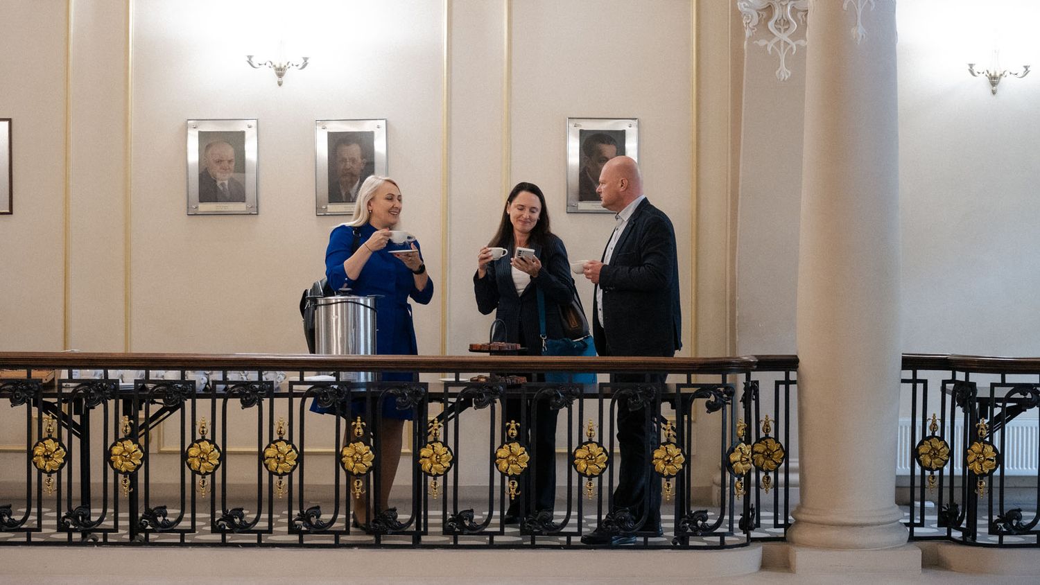 Three people chat and hold drinks on a wrought-iron balcony beneath framed portraits in an elegant, columned hall.